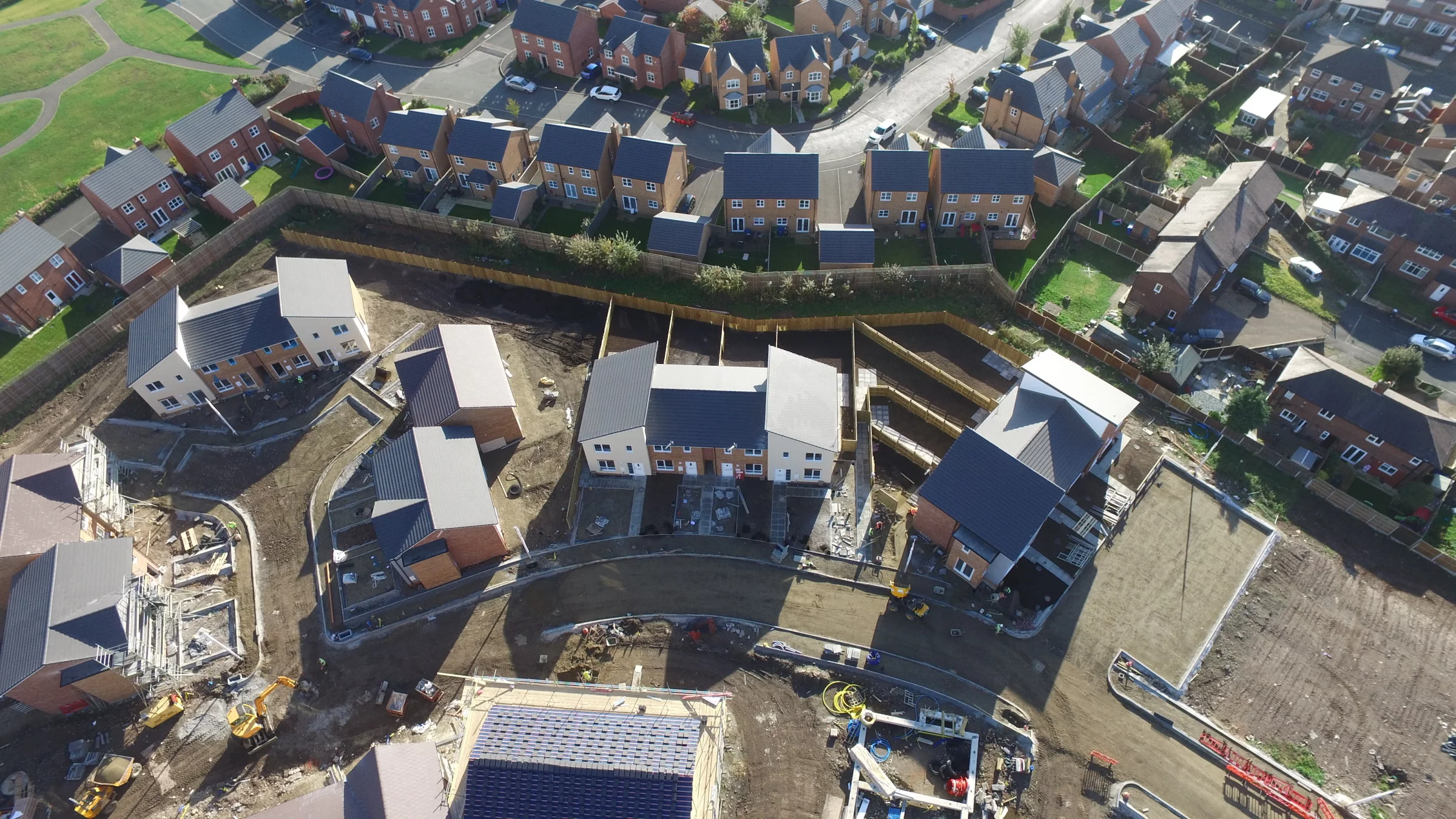 Aerial view of a new housing estate: finished terraced houses with fenced gardens above a construction site with diggers and machinery.