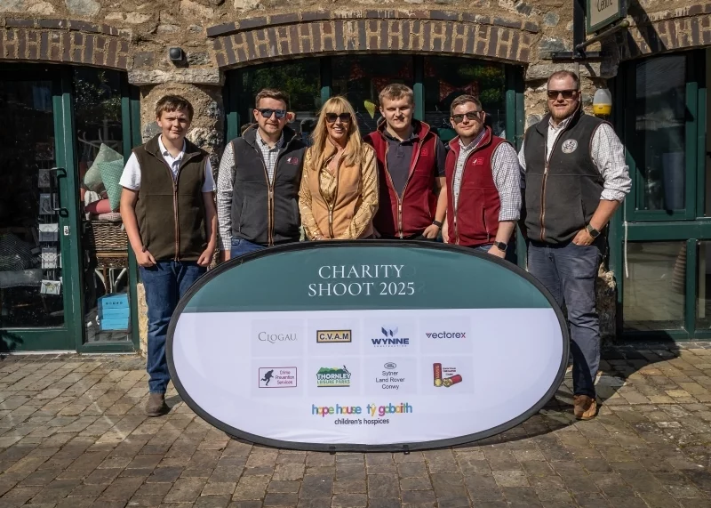 Six people in outdoor vests stand behind an oval sign reading CHARITY SHOOT 2025 with sponsor logos, outside a stone building.