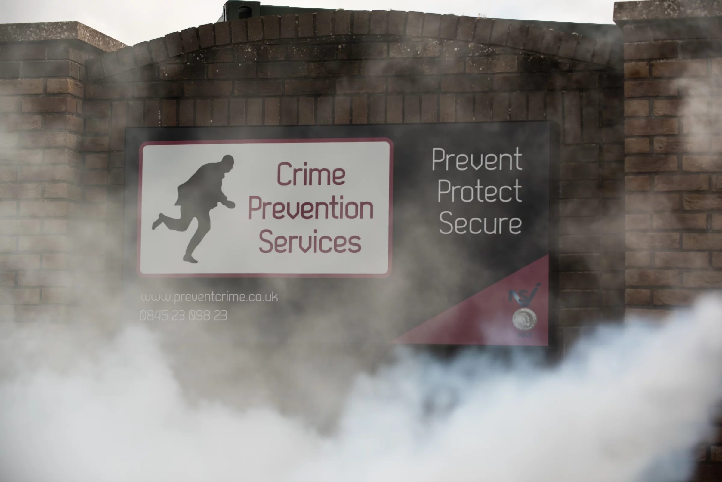 Brick sign reading Crime Prevention Services with thief silhouette and words Prevent, Protect, Secure seen through fog.
