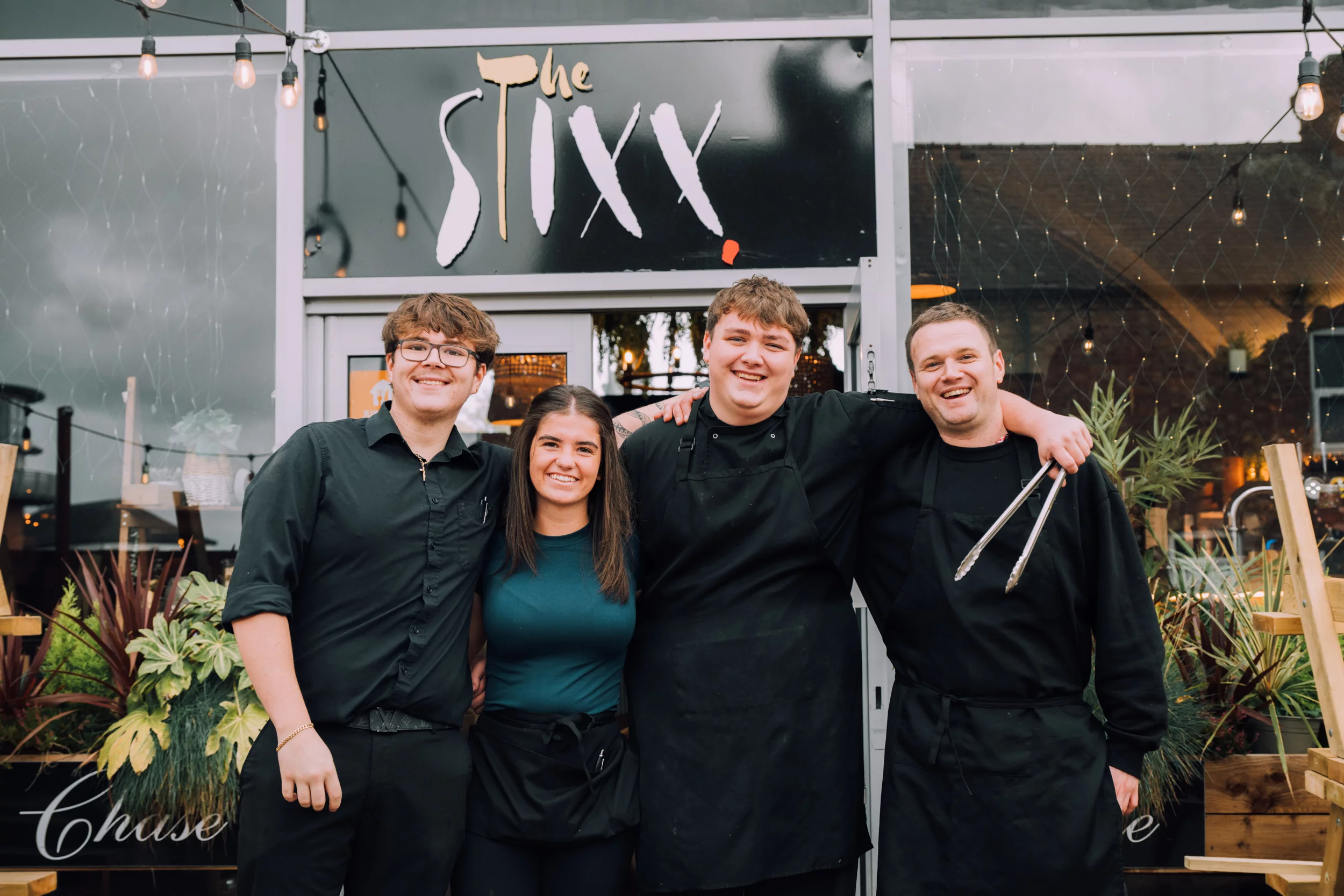 Four smiling restaurant staff in black uniforms arm-in-arm under The SIXX sign outside a lit café, one holding tongs.