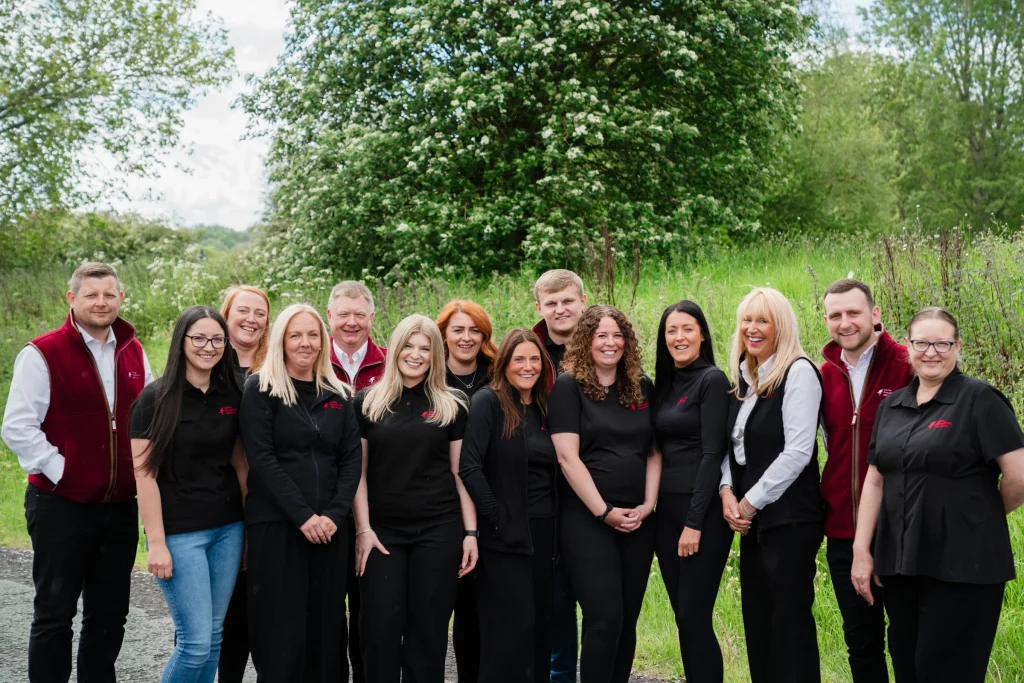 Group of fourteen smiling staff in black tops and red vests standing outdoors before a flowering tree.