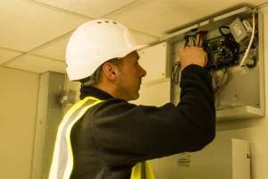 Construction worker in white hard hat and high-visibility vest wiring an open electrical control panel.