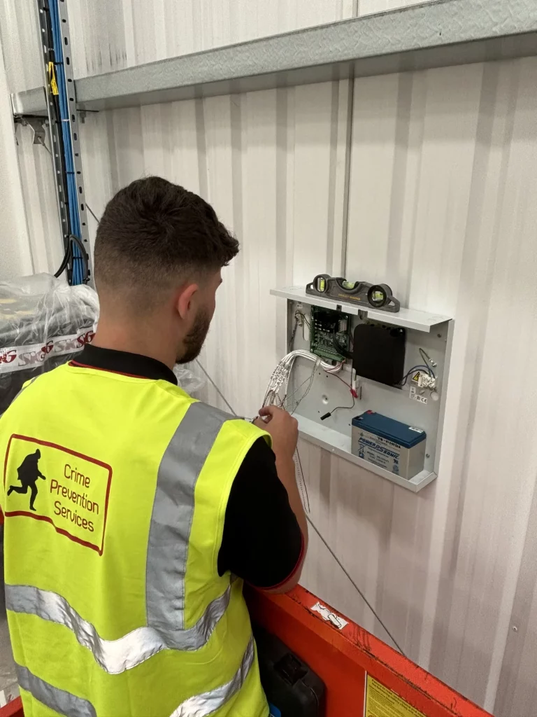 Worker in high-vis vest labelled Crime Prevention wiring an open wall-mounted control panel with a spirit level on top.