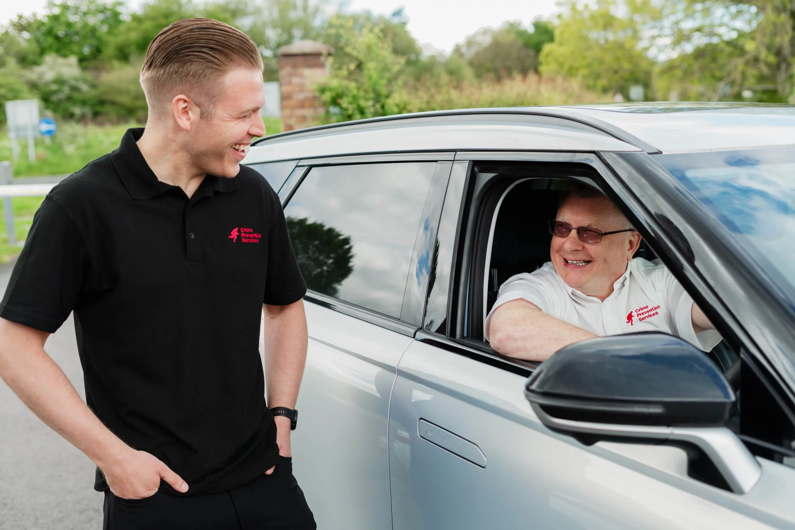 Young man in black polo smiling at an older man in a white shirt leaning from a silver car window.