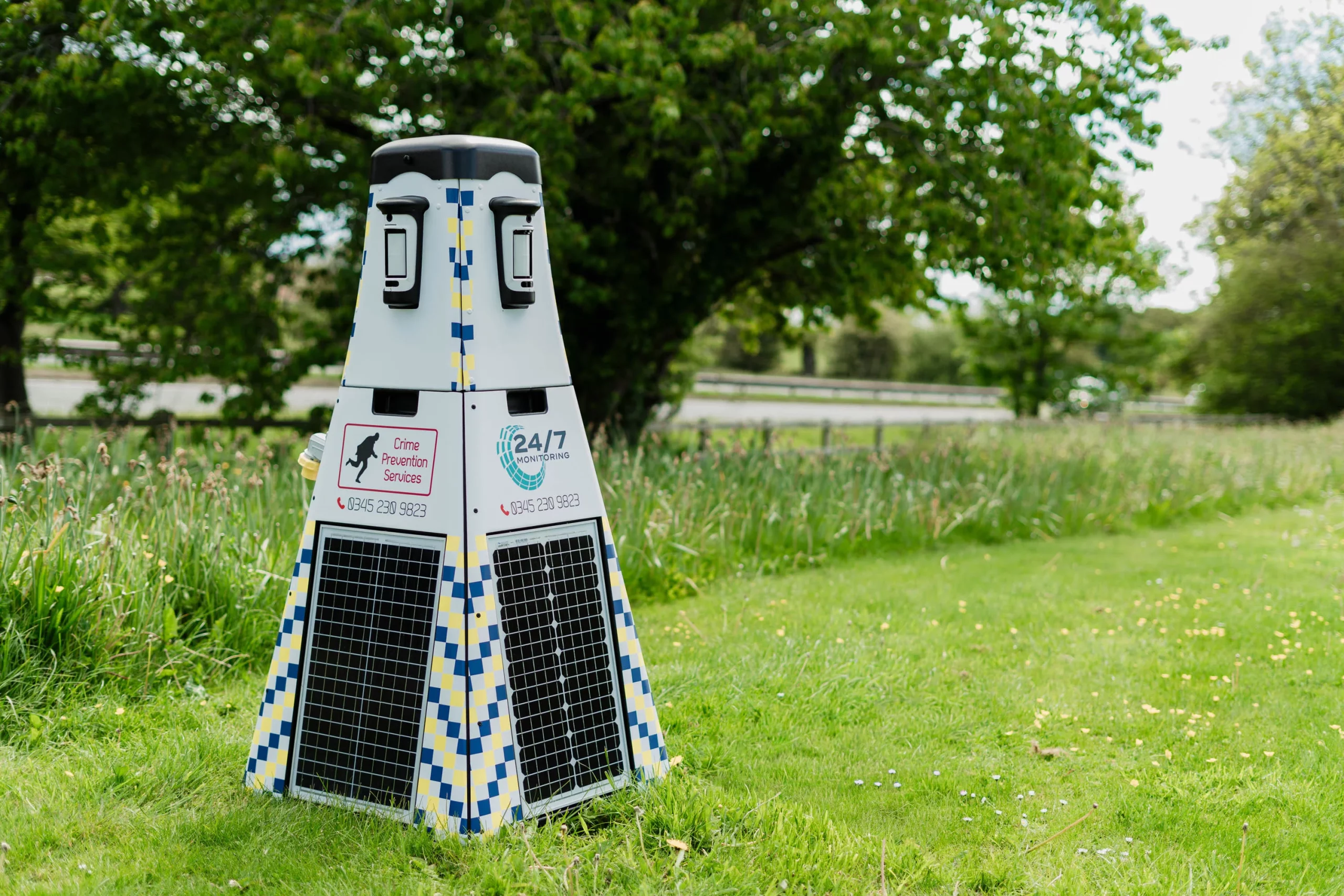 Pyramidal surveillance unit with solar panels and blue-yellow checkered markings labelled 24/7 Monitoring on a grassy verge