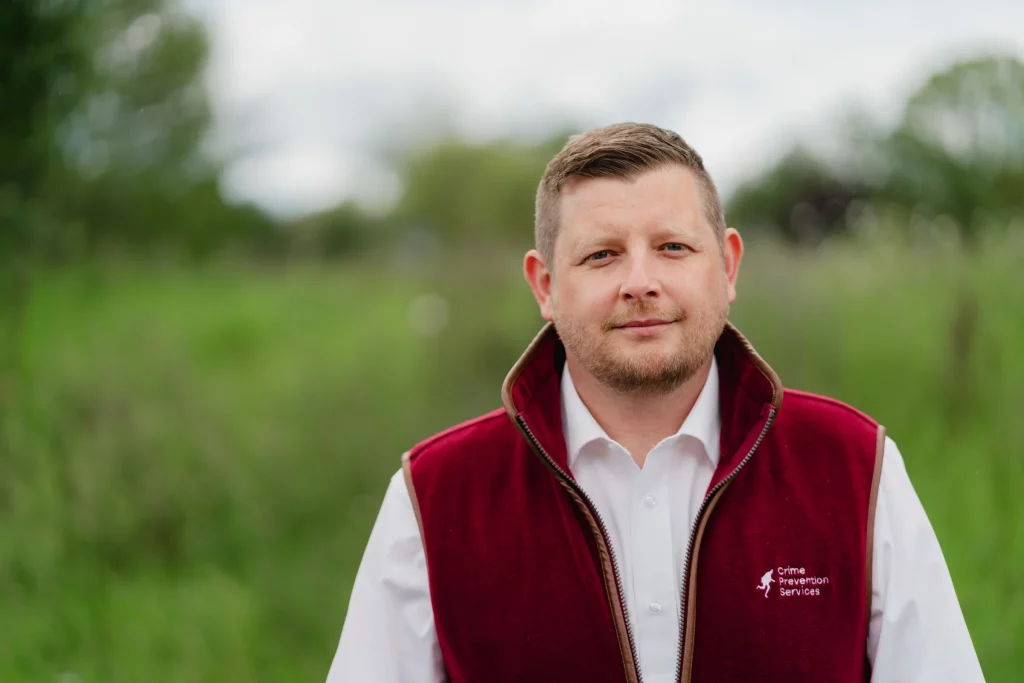 Man with short hair and light beard wearing a maroon vest embroidered Crime Prevention Services, standing outdoors with blurred greenery background.