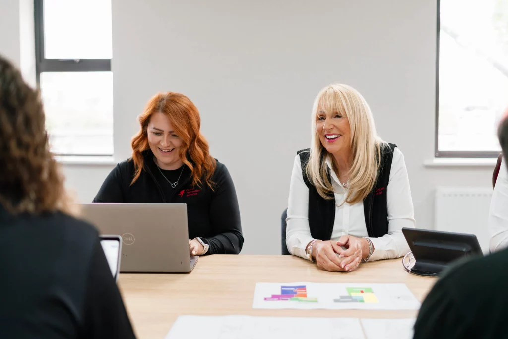 Two women at a meeting smiling, a red-haired woman using a laptop and a blonde woman beside her with hands clasped.