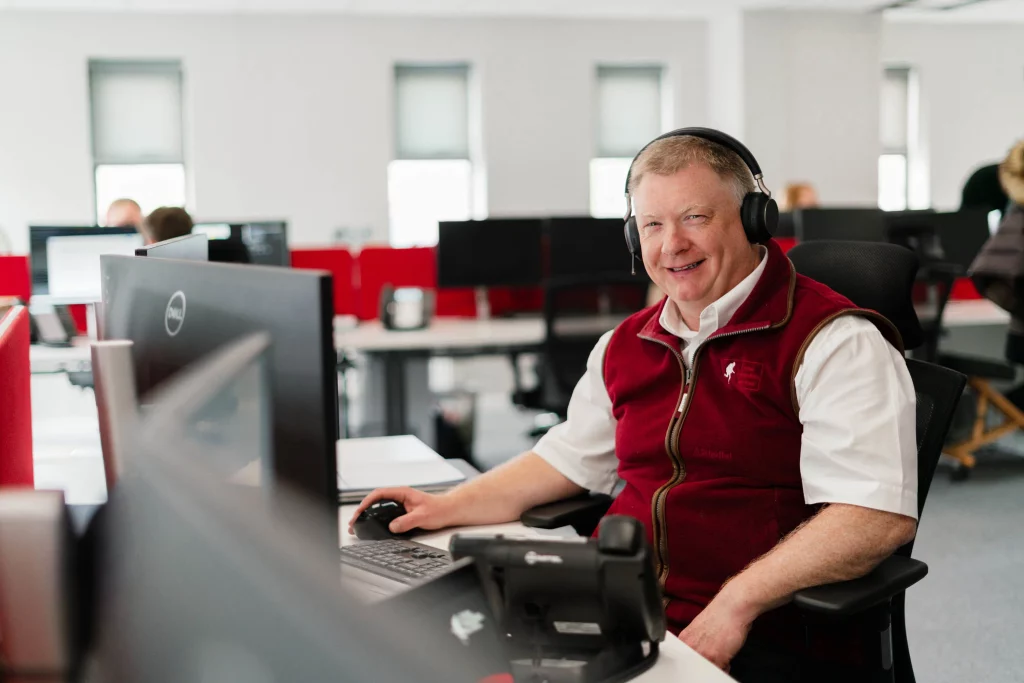 Smiling man wearing a headset and red zip-up vest seated at a computer in an open-plan office.