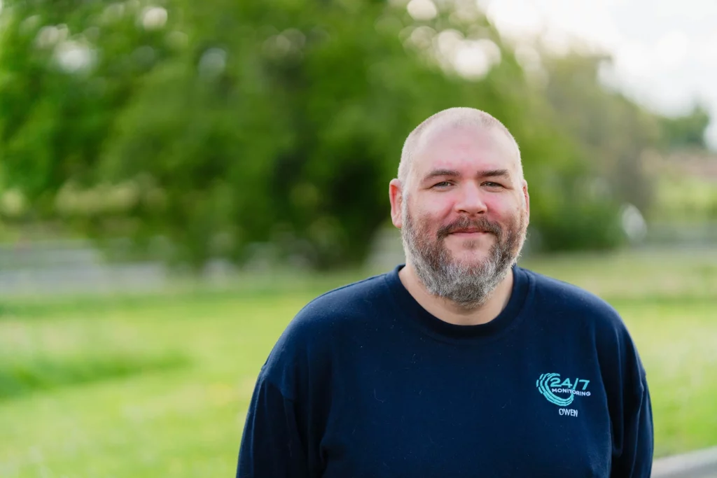Smiling bearded man with shaved head in a navy shirt with 24/7 logo, standing outdoors with blurred green trees