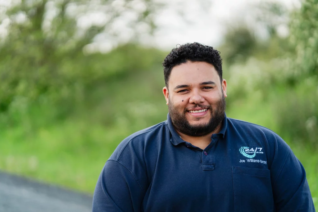 Smiling bearded man with short curly hair wearing a navy polo branded "24/7 Monitoring", blurred green background.