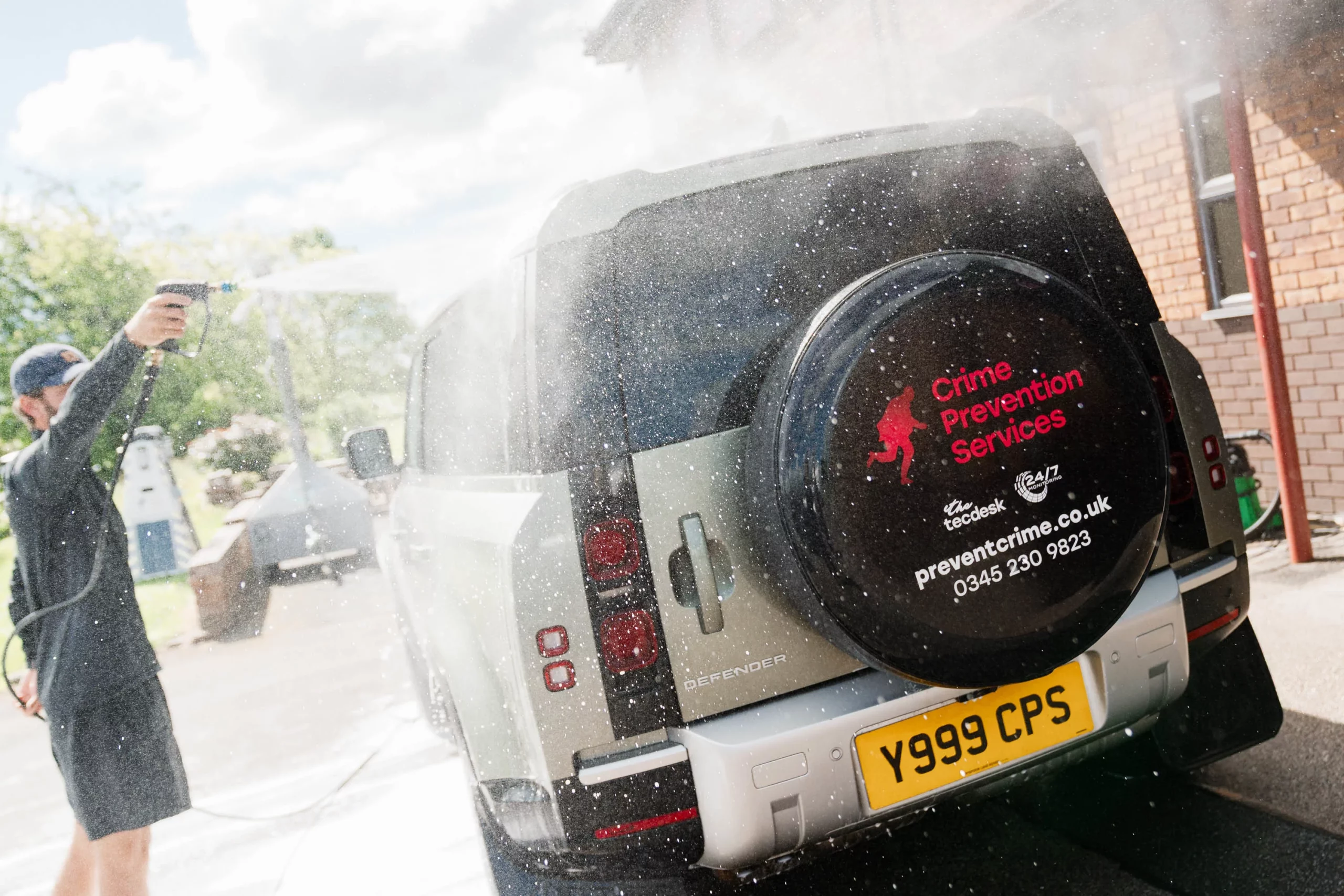 Person pressure-washing a silver SUV rear, water spray over a spare tyre cover labelled Crime Prevention Services.