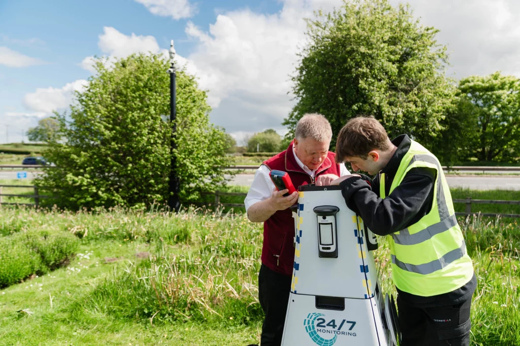 Two technicians, one in a hi-vis vest, inspecting a white 24/7 monitoring unit beside a grassy roadside with trees.