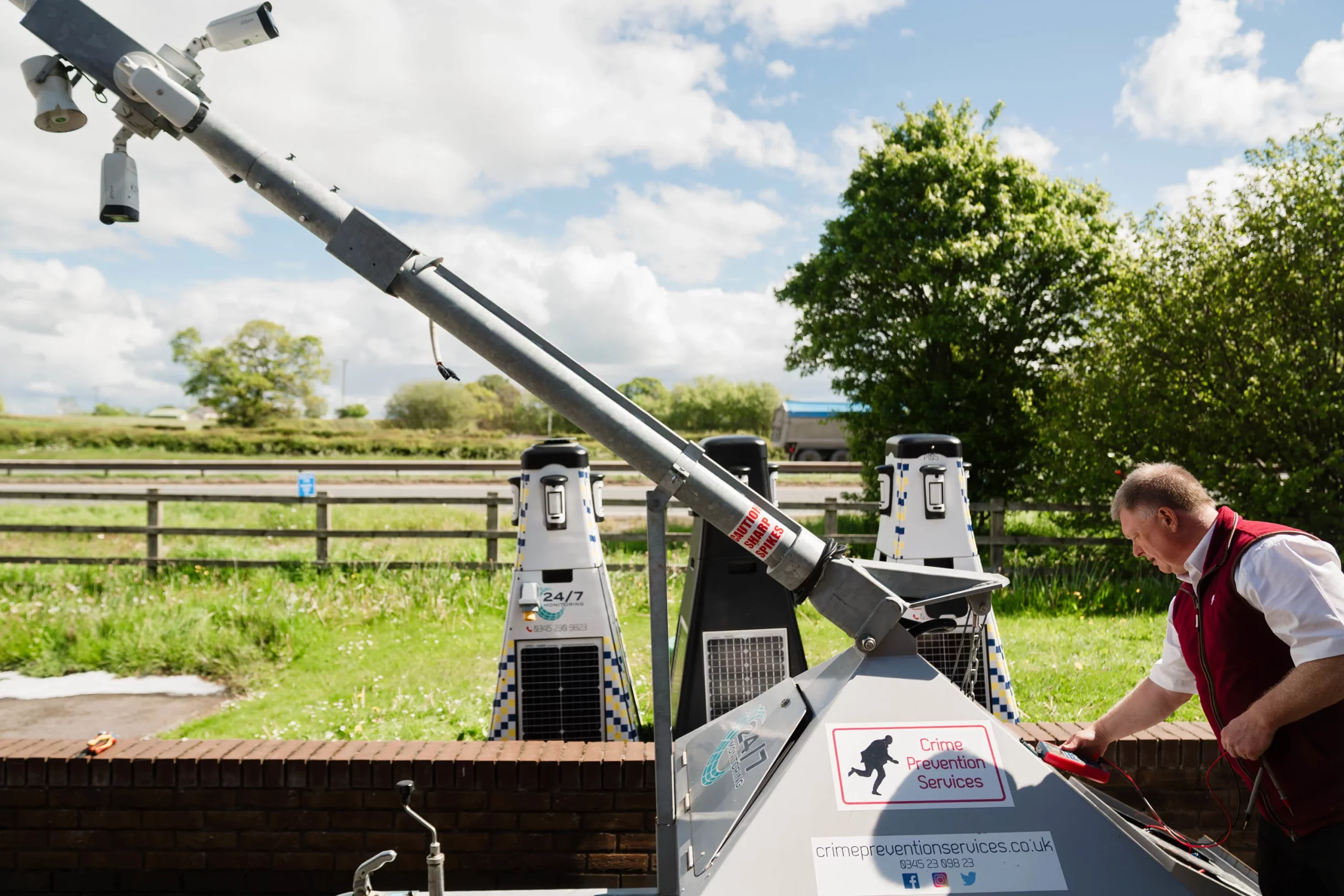 Tilted pole with CCTV cameras and mobile crime-prevention units, a man adjusting equipment at a roadside.