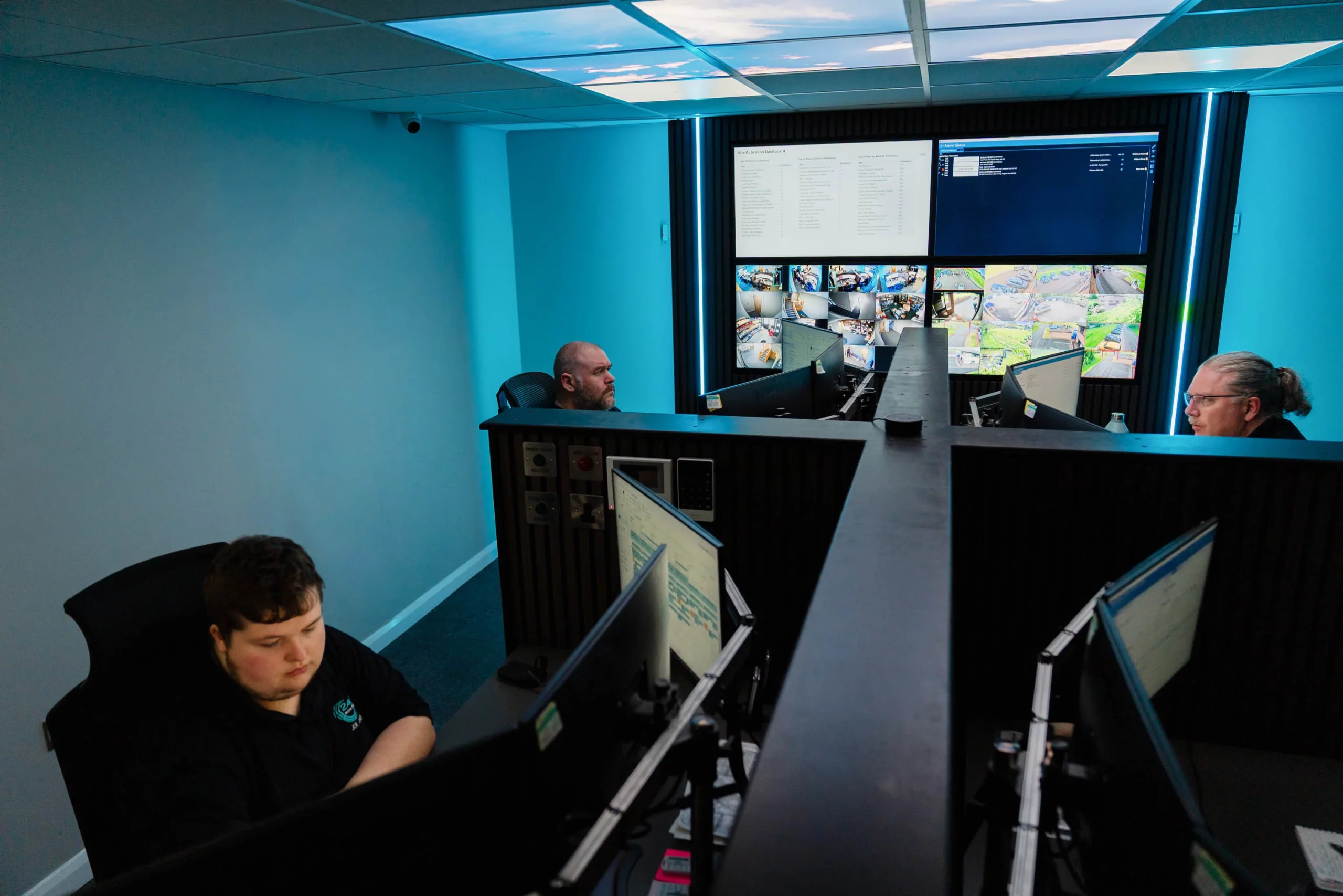 Control room with three operators at desks facing multiple monitors and a large wall of CCTV feeds under blue lighting.