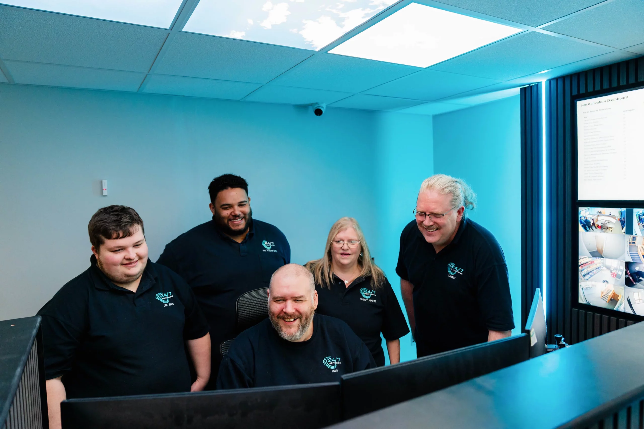 Five colleagues in matching dark polo shirts smiling around computer monitors in a blue-lit control room with CCTV screens.