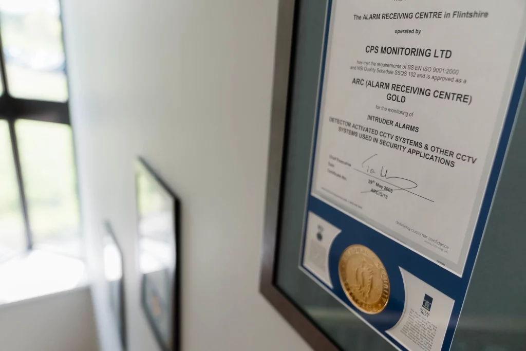 Framed certificate with gold seal for CPS Monitoring Ltd (ARC Gold) on a wall beside a stairwell.