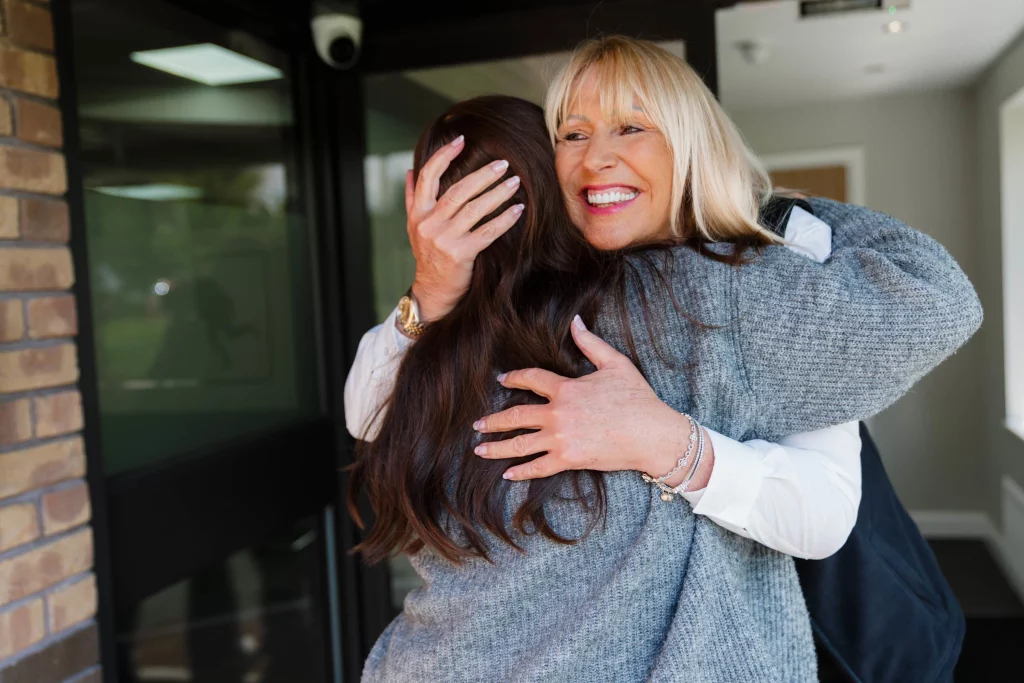 Blonde woman smiling broadly as she embraces another woman with long brown hair in a warm hug at a doorway