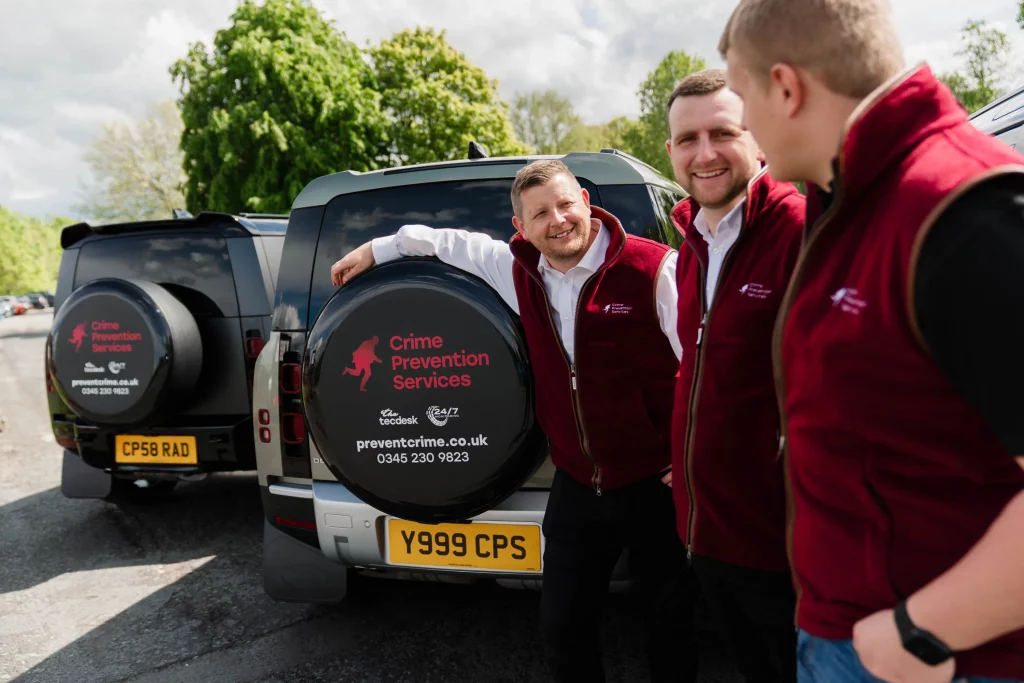 Three men in burgundy Crime Prevention Services vests chatting beside branded SUVs with spare-tyre covers in a car park