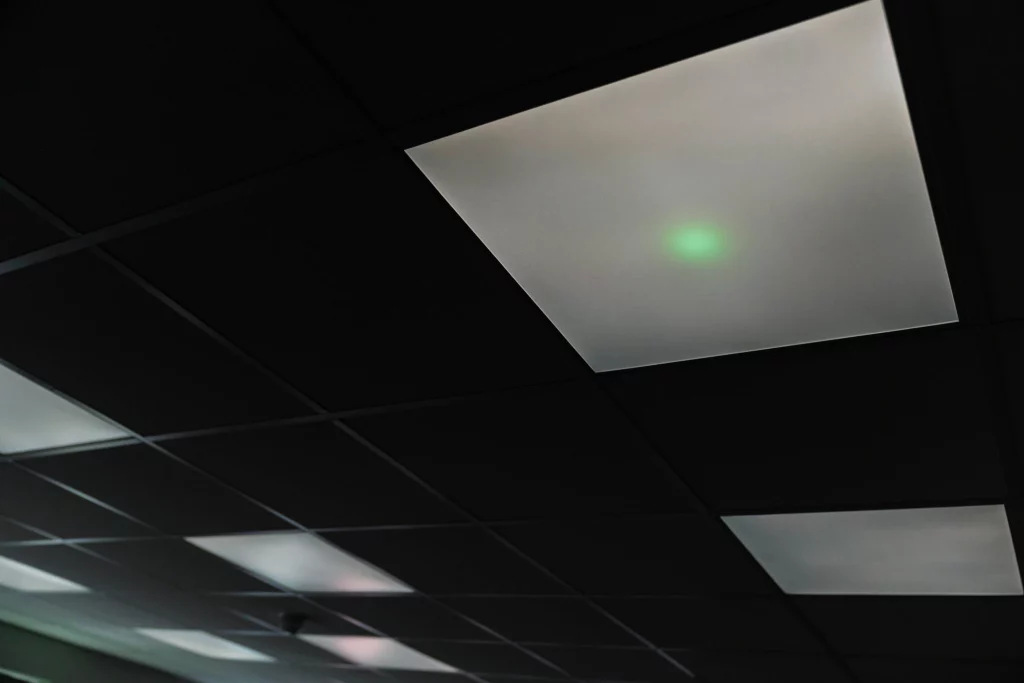 Drop ceiling with white recessed light panels against dark tiles, one panel showing a small green glowing spot.