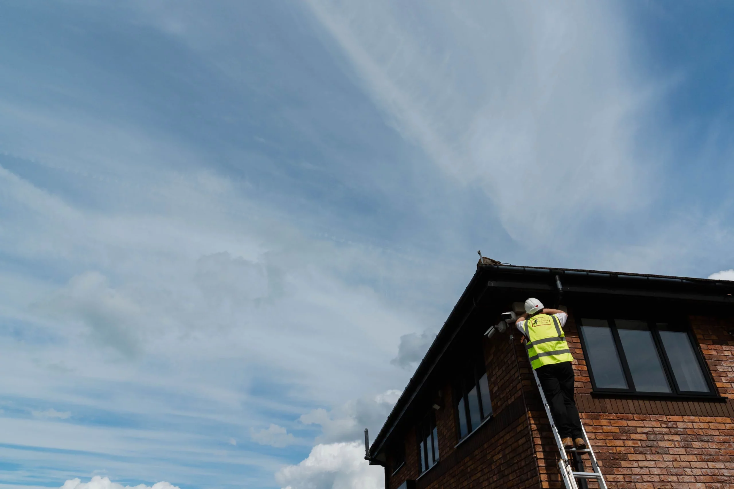 Worker in a hi-vis vest and hard hat on a ladder fitting a security camera under the house eaves against a blue sky.