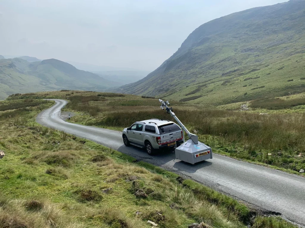 White pickup towing a box trailer with mounted equipment parked on a narrow winding road through misty green hills.