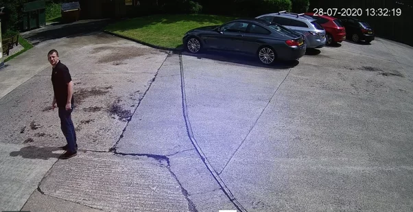 Man in dark shirt and jeans stands on cracked concrete car park looking over his shoulder at the camera, parked cars and timestamp visible.