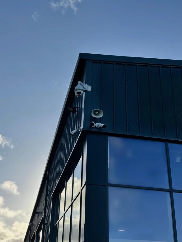 Corner of a dark-clad building with reflective windows, security cameras and a wall-mounted speaker against a blue sky.