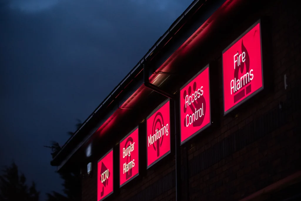 Illuminated red square signs on a brick building at dusk reading Access Control and Fire Alarms.